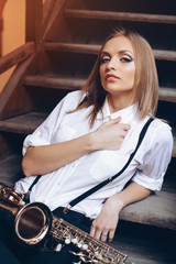 Fototapeta na wymiar Young attractive girl sitting on steps in white shirt with a saxophone - outdoor. Sexy young woman with sax looking at camera