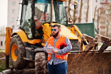 Brutal beard worker man suit construction worker in safety orange helmet, sunglasses against traktor with plan paper at hands.