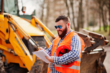 Brutal beard worker man suit construction worker in safety orange helmet, sunglasses against traktor with plan paper at hands.