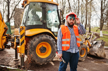 Brutal beard worker man suit construction worker in safety orange helmet, sunglasses against traktor with adjustable wrench at hand.