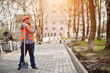 Fototapeta na wymiar Portrait of brutal beard worker man suit construction worker in safety orange helmet against pavement with shovel in hand.