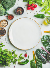 Fresh raw greens, unprocessed vegetables and grains over light grey marble kitchen countertop, wtite plate in center, top view, copy space. Healthy, clean eating, vegan, detox, dieting food concept