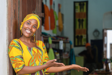 Smiling black African woman in traditional clothes wishing welcome to customers in front of her...