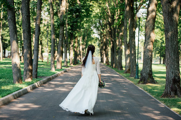 Beautiful young smiling brunette bride in wedding dress with bouquet of flowers in hands goes in the park outdoors