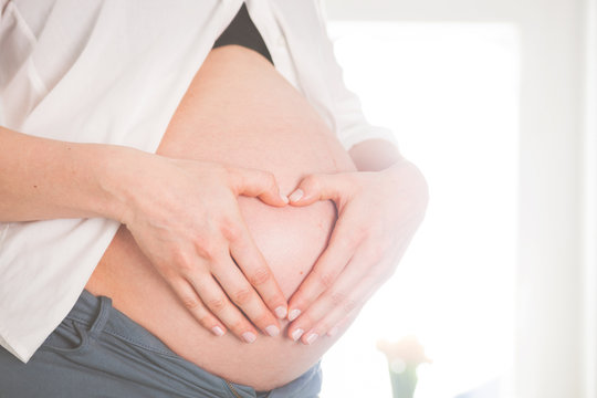Pregnant girl holding hands in heart shape on belly at home