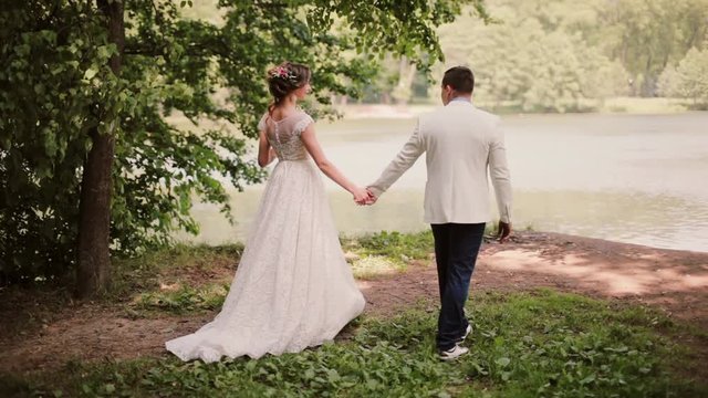 Beautiful couple share wedding day. They walk in park near river holding hands. Bride and handsome groom. Steadicam shot