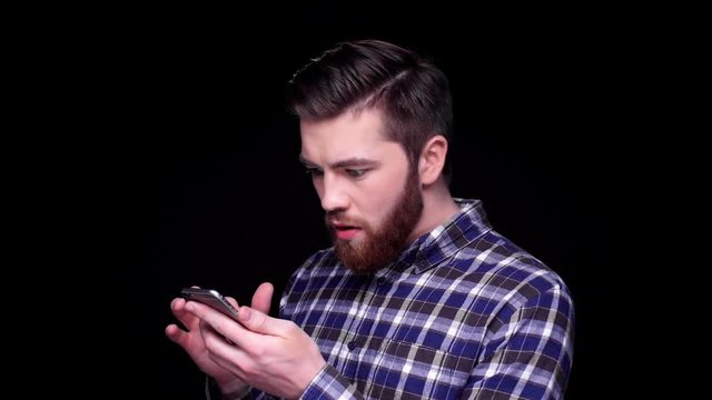 Young excited bearded man receiving good news on smartphone isolated over black 