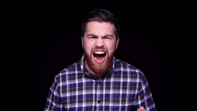 Angry furious young bearded man shouting at camera and pointing finger isolated over black 