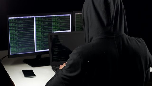 Hacker in hood cracking code using laptop and computers from his dark hacker room