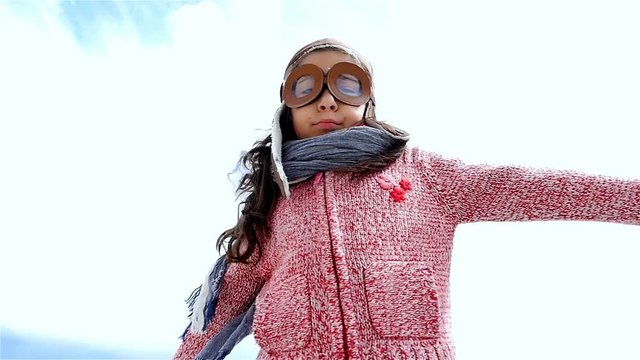 Little girl with pilot hat playing spreading arms like an airplane against blue sky, slow motion