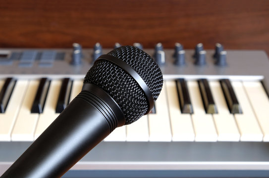 Black vocal microphone close up against electronic synthesizer keyboard with many control knobs in silver plastic body as background