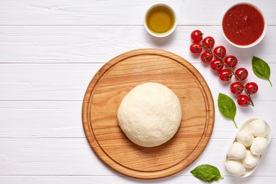 Raw pizza dough with baking ingredients on cutting board: dough, mozzarella, tomatoes sauce, basil, olive oil, cheese, spices. Italian margherita on wooden table. Italian pizza margarita