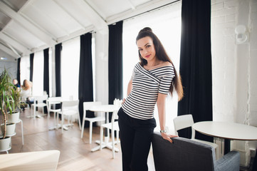 Obraz premium Sexy beautiful woman wearing stylish striped blouse shirt. Designer of fashion clothes posing and looking at the camera in a cafe with huge windows behind her.