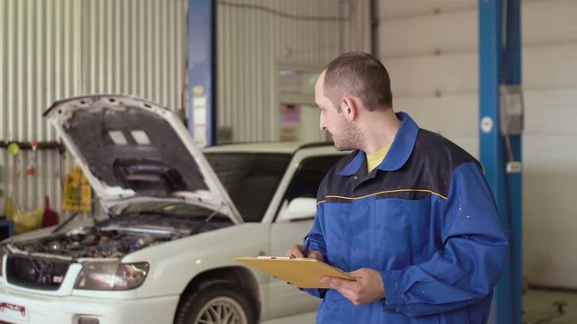 Portrait of a car mechanic writing in a note pad clipboard behind car with opened hood in a repair car service