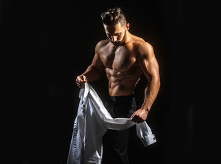 Man undresses, takes off his white shirt. Young guy with beautiful bare naked sports torso. Man wearing shirt. Sexy young  muscular man in shirt on a dark background at morning