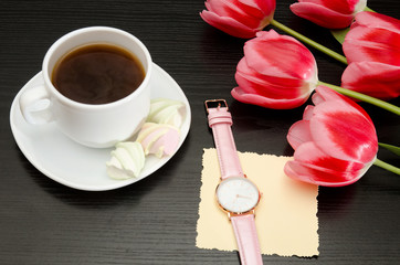 Mug with coffee and marshmallow, net postcard, watch, pink tulips. Black table. Top view