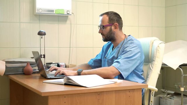 Young, male doctor working on laptop sitting by table in office
