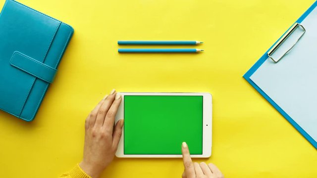 Woman using her white tablet device with green screen, scrolling news, tapping on photos. Colorful desk background. Yellow table. Top view. Chroma key