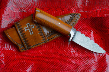 A Christian themed leather sheath and fixed blade knife sitting atop a man made cobra skin dyed red