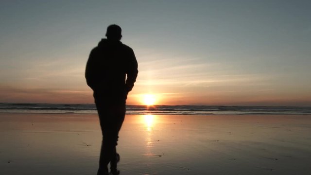 Silhouetted man casually walks out on low tide beach to enjoy the peaceful sunset over the Pacific Ocean.