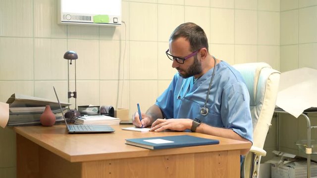 Male doctor writing rx prescription sitting by table in office in hospital 
