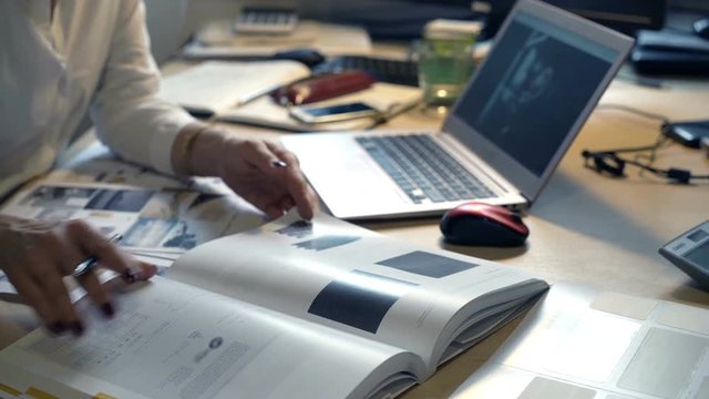 Female designer with laptop checking products in book sitting in office
