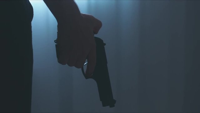 DOLLY IN Silhouette of male criminal pulling back the hammer of a pistol near the window. 4K UHD RAW edited footage