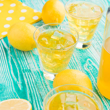 lemonade or limoncello in glasses with ice cubes, sherbet glass with ice cubes, bottle with drink, lemon fruits on turquoise colored wooden table
