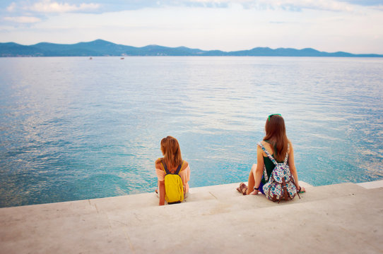 Two young girls with backpacks sitting on the stone stairs near the sea shore looking at the hill range in the distance. Town of Zadar, Croatia