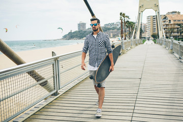 Handsome man in summer clothes walking on pier with skateboard in hand