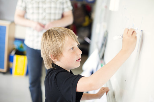 Side view of boy writing on whiteboard while teacher standing in background at classroom