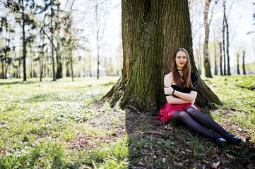 Portrait of girl with bright make up with red lips, black choker necklace on her neck and red leather skirt sitting near tree at park.