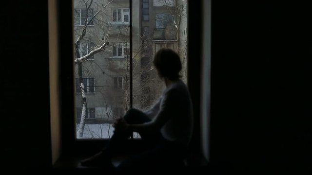 Depressed woman sitting on the sill and looking out the window, broken-hearted