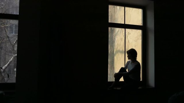 Silhouette of sad woman sitting on the sill and looking out the window