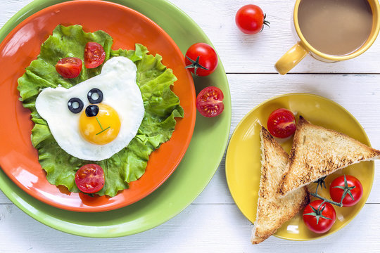 Funny Breakfast with bear-shaped fried egg, toast, cherry tomato, lettuce on colored plates and coffee.