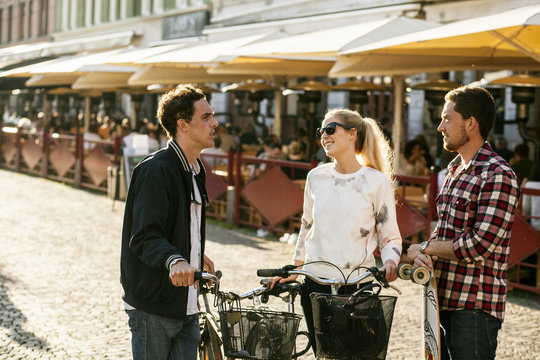 Male and female friends with bicycles and skateboard conversing by sidewalk cafe