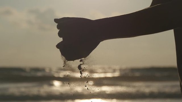 Silhouette of female hand pouring sea sand through her fingers at sunset against an ocean background. Arm of young woman with sand strewed or falling from it. Grit drizzling from fist of girl. Closeup