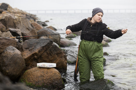 Man repairing fishing rod while standing in lake by rocks