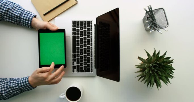 Man working on laptop and black tablet device with green screen on wite office table. Using touch screen. Top view. Chroma key