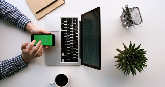 Top view of man using laptop and black smart phone with green screen on wite office table. Scrolling pages, tapping on touch screen of cell phone. Chroma key