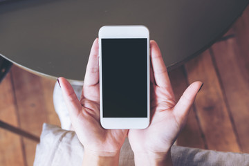 Mockup image of hands holding white mobile phone with blank black screen on pillow with wooden floor background