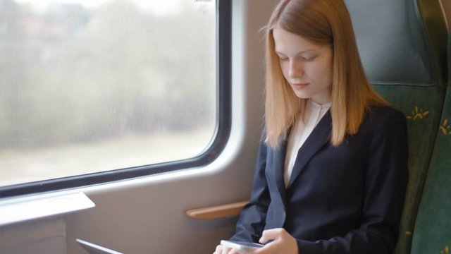Young Beautiful Business Lady is Sitting by the Window in the Train and Working on her Laptop and Looking at the Phone