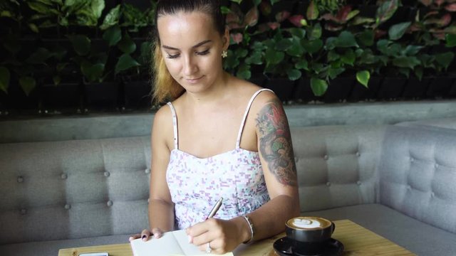 Young prety woman with tattoo writing in her journal, diary. Caucasian model with long hair. Cup of coffee and smartphone on the table,