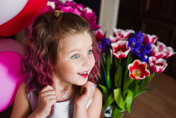 Little beautiful sweet girl with pink curly hair in a silver dress in red tulips and with multi-colored balls smiling and posing. Happy childhood like a princess