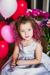 Fototapeta na wymiar Little beautiful sweet girl with pink curly hair in a silver dress in red tulips and with multi-colored balls smiling and posing. Happy childhood like a princess