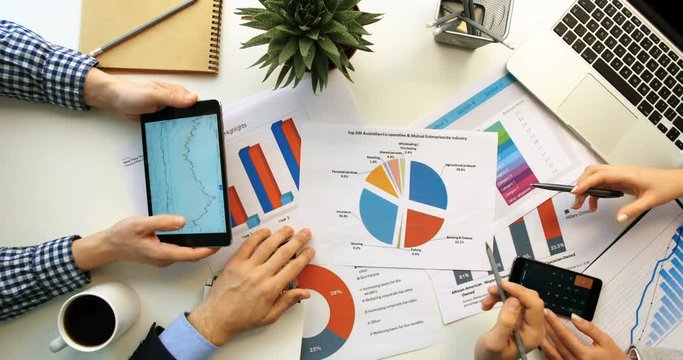 Business team working together at office desk and checking a financial report, men using tablet and charts and woman using smartphone. Hands top view.