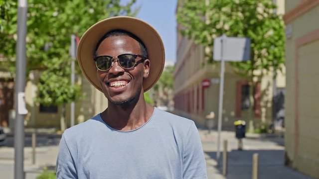 Hipster happy young black man swiftly walks around the summer city wearing classic vintage sunglasses and a trendy hat, laughs and smiles into the camera enthusiastic and optimistic