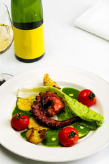 Seafood dish with fried octopus tentacles and vegetables with a bottle and a glass of wine on a white tablecloth background, studio light