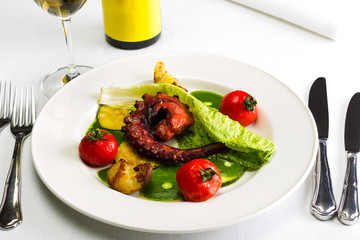 Seafood dish with fried octopus tentacles and vegetables with a bottle and a glass of wine on a white tablecloth background, studio light