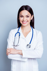 Vertical portrait of charming smiling young female doctor in uniform on gray backgroung standing with crossed hands